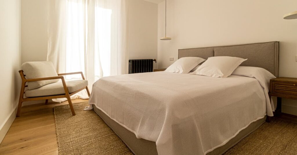 Dormitorio minimalista con cama doble, cabecero tapizado en gris y luz natural entrando por la ventana